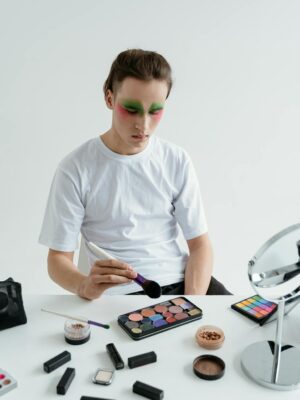 Man in white shirt applying makeup with eyeshadow and brushes on table.